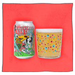 Beavertown Brewery Lazer Crush - Alcohol Free IPA and Beavertown Psychedelic Skull Tumbler glass filled with beer in front of a pink background. Beer glass is clear glass that has red, blue, pink and yellow skulls and bones speckled all over.